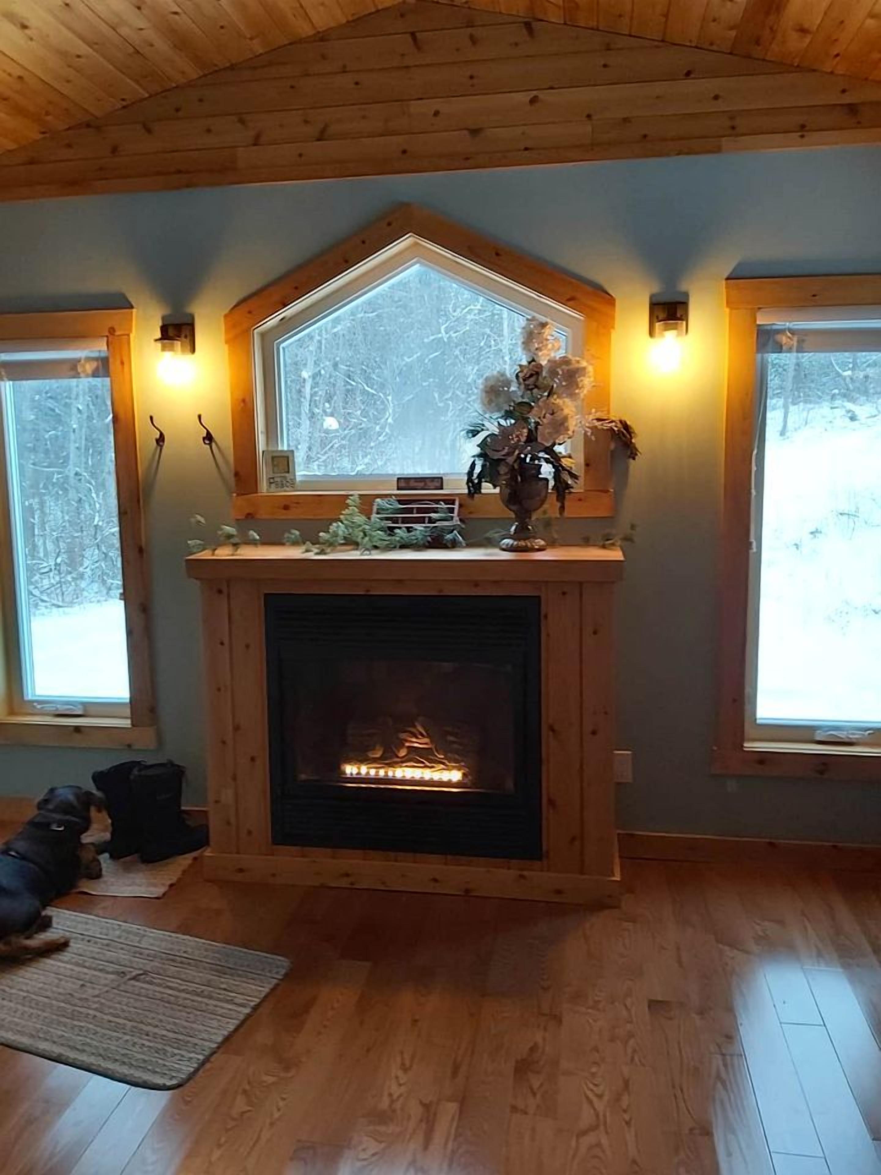 Une pièce dotée d’une cheminée, d’un plafond en bois et de grandes fenêtres offrant une vue sur les arbres.