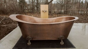 Une baignoire autoportante, soutenue par une structure en bois, posée sur un tapis texturé à l’extérieur.