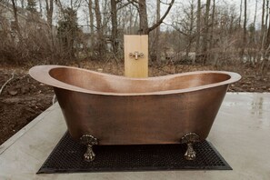A freestanding bathtub with a wooden support, placed on a textured mat outdoors.