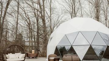 A geodesic dome structure with a transparent window, surrounded by outdoor furniture including a wicker chair, a wooden tub, and two stools.