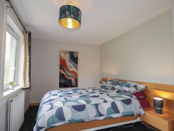 Un dormitorio con una cama, una mesita de noche con lámpara, una ventana con cortinas y una obra de arte colgada en la pared.