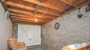 Une pièce avec un plafond en bois, un jeu de fléchettes, une porte de garage blanche, des meubles en osier et un mur en briques.