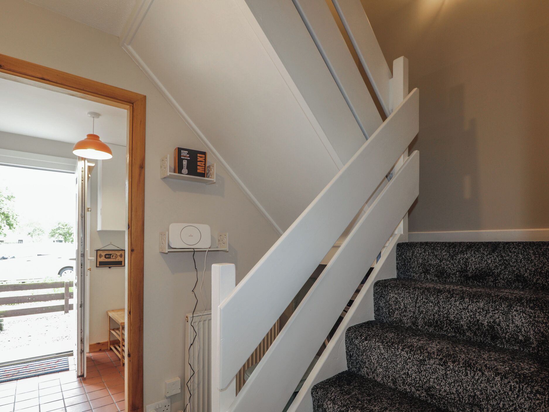 A staircase with a white handrail and patterned steps, a door leading outside, and a shelf with books and a device on the wall.