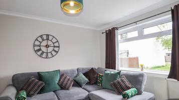 A grey sectional sofa with multiple cushions, a wall clock, a window with curtains, and a pendant light fixture.