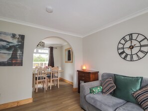 A living room with a grey sofa, a wooden dining table with chairs, a wall clock, and a painting. - Taigh Ban (Nairn)