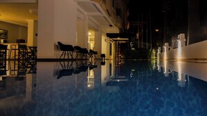 Vista nocturna de una piscina con sillas y mesas, entrada a un edificio y una calle iluminada.
