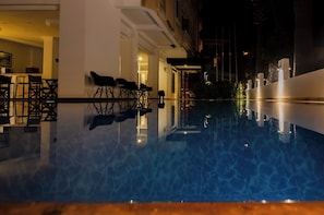 Szenenbild eines Schwimmbeckens bei Nacht, mit Liegestühlen und Tischen, einem Gebäudeeingang und einer beleuchteten Straße.