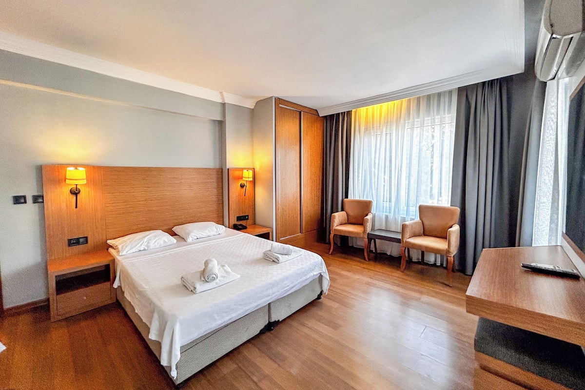 A hotel room with a large bed, two bedside tables, a desk, two chairs, and a window with curtains.