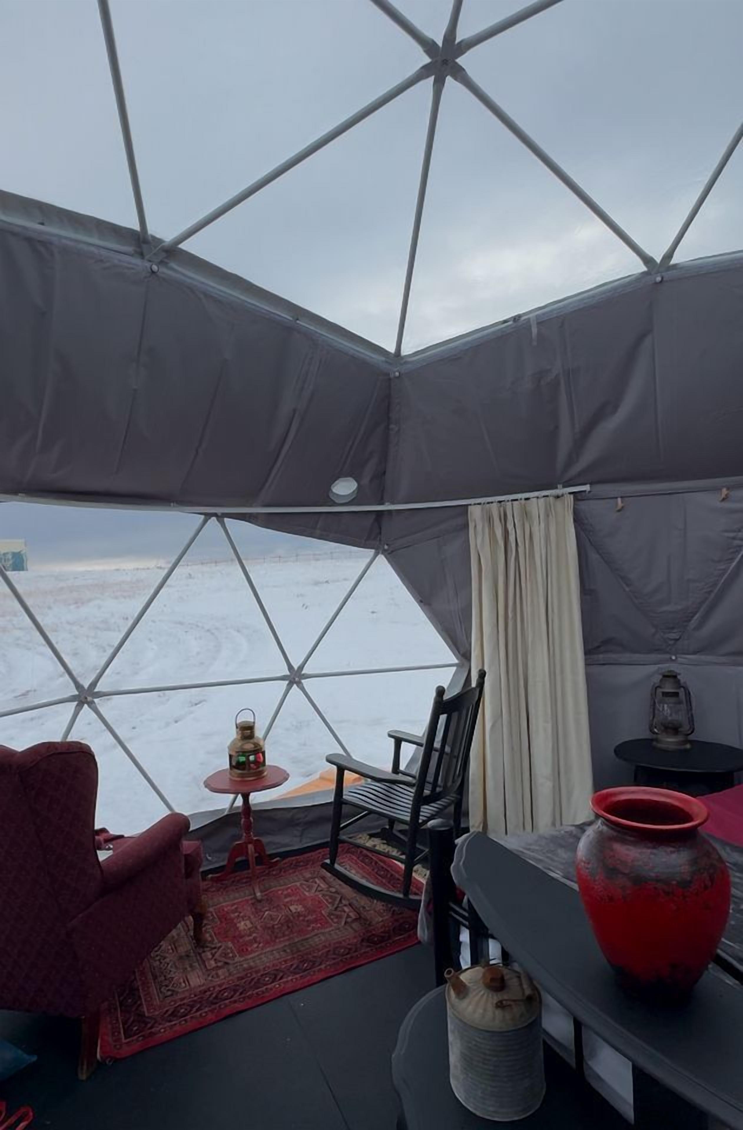 Een tent met uitzicht op een besneeuwd landschap, een rode stoel, een klein tafeltje met een fles en een lantaarn.