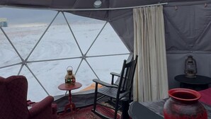 A tent with a view of a snowy landscape, a red chair, a small table with a bottle, and a lantern. - Cozy Geodesic Glamping Dome on a Canadian Ranch with Horses, Perfect for a Weekend of Wellness (Redcliff)