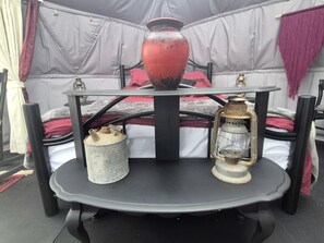 A tent with a bed, a table with a vase and lantern, and a metal container.