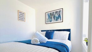 Una cama bien hecha con una colcha azul y blanca, una cita inspiradora en un marco en la pared, y un cuadro encima.