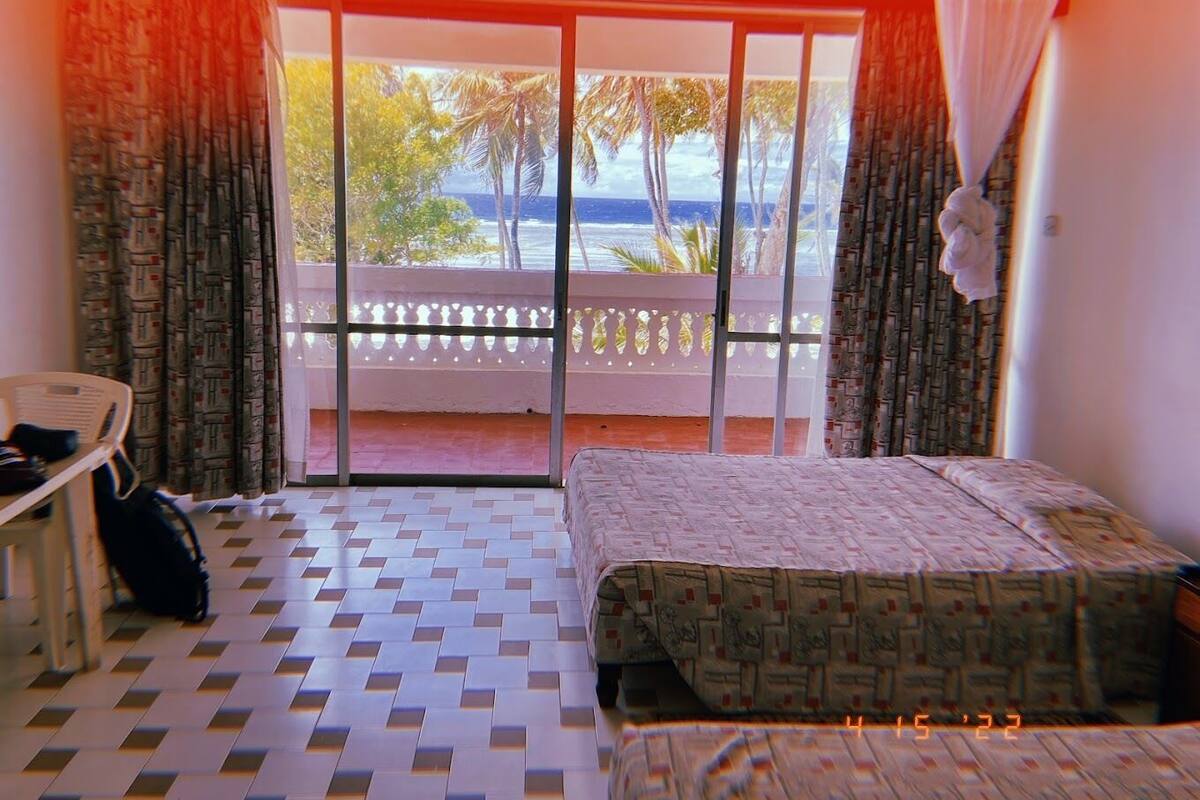 A hotel room with a bed, a chair, a suitcase, a balcony with a view of the sea and palm trees, and a patterned floor.