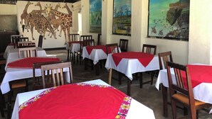 A restaurant with tables covered in red and white tablecloths, wooden chairs, and walls adorned with paintings. - Twiga Lodge Tiwi (Tiwi)
