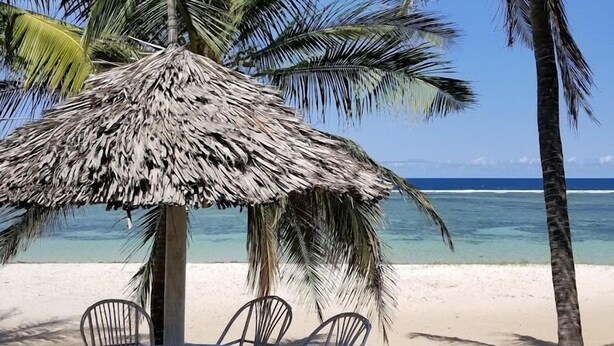 A beachside table with chairs under a thatched umbrella, palm trees, and a view of the ocean. - Twiga Lodge Tiwi (Tiwi)