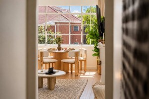A dining area with a round table, four chairs, a vase with flowers, and a view of a brick building through large windows. - Two-Bedroom Flat with Parking Near Bondi Beach (Bondi)