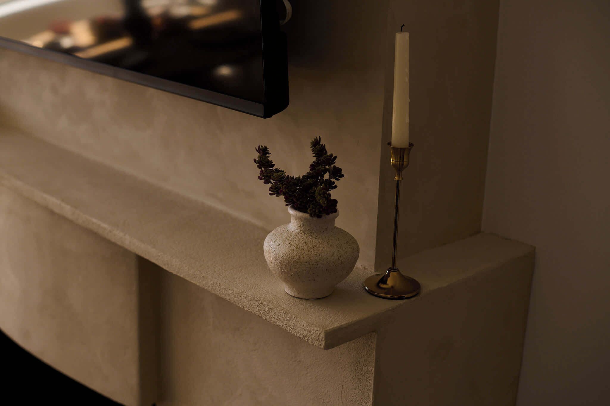 Un florero pequeño y redondo, con textura, con una planta sobre una superficie plana, al lado de un candelabro alto y delgado con una vela blanca.