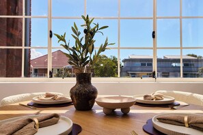 Una mesa de comedor con platos, tazones y un florero con una planta, frente a una ventana con vistas a edificios.