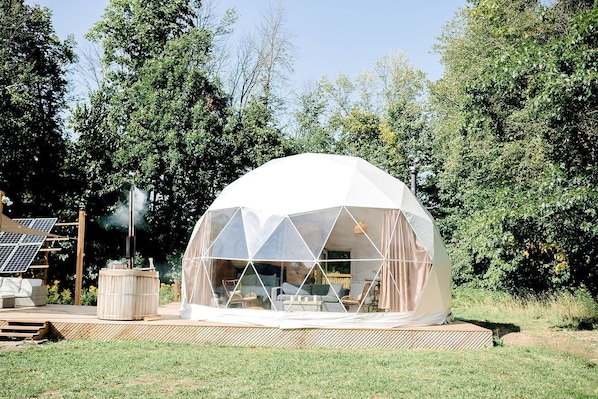 Une structure en forme de dôme géodésique dotée d’un toit transparent, entourée d’une terrasse en bois, avec des panneaux solaires en arrière-plan.