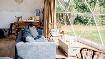 Un intérieur chaleureux avec une grande fenêtre, un canapé, un fauteuil en osier, une petite table avec des bougies et une bouteille d’eau.
