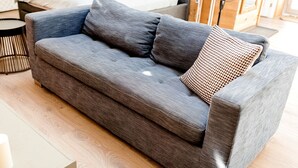 A grey sofa with a woven pillow, a small side table, and a fan in a room with a wooden floor. - Unique Off-Grid Dome Escape with Hot Tub & Forest Views in Ontario, Canada (Marmora)