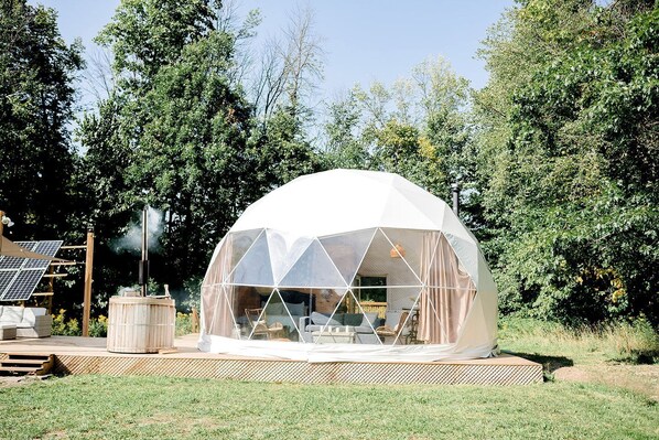 Estructura en forma de cúpula geodésica con techo transparente, rodeada de una terraza de madera, y paneles solares al fondo.