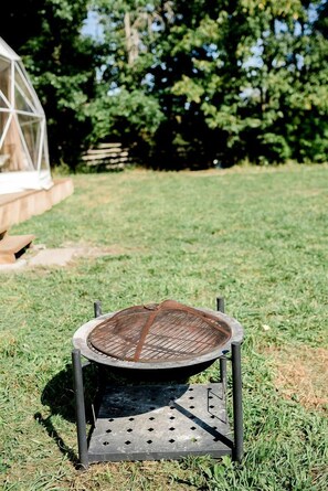 Un barbecue rond en métal, doté d’une grille supérieure en treillis, posé sur un support métallique à base perforée, dans un espace extérieur verdoyant, avec une clôture en bois et des arbres en arrière-plan.