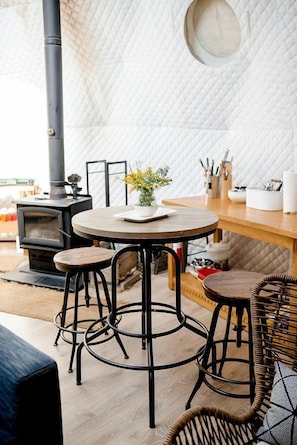 A cozy room with a round wooden table, two stools, a wicker chair, a white quilted wall, and a wood-burning stove.