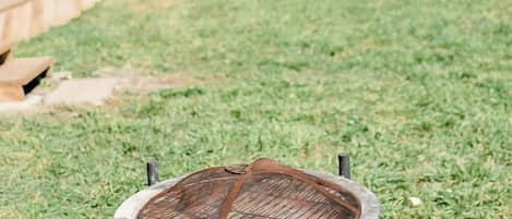A round metal grill with a mesh top, set on a metal stand with a perforated base, in a grassy outdoor area with a wooden fence and trees in the background.