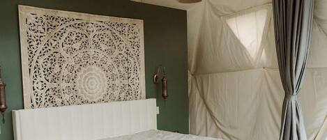 Un dormitorio con cama, lámpara de fibras naturales, panel decorativo en la pared y vistas a través de una ventana con cortinas.