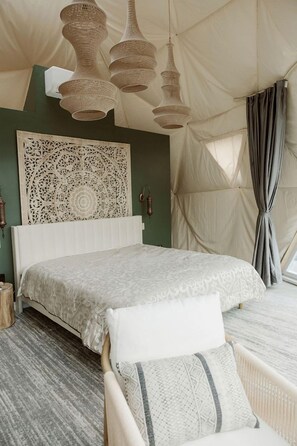 A bedroom with a bed, a woven light fixture, a decorative wall panel, and a view through a window with curtains.