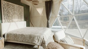 A bedroom with a bed, a chair, a large window, and a decorative wall piece. - Peaceful Boho Dome Escape with Open Field Views in Ontario, Canada (Marmora)