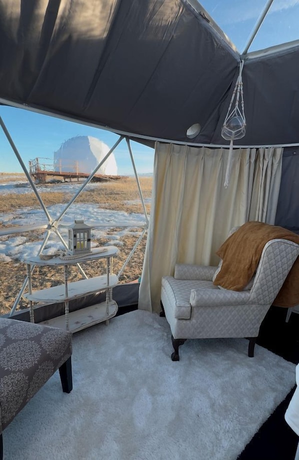 A tent with a view of a snowy landscape, a cushioned chair, a small table with a lantern, and a white carpet.