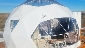 A geodesic dome structure with a transparent window section, wooden deck, and a clear sky.