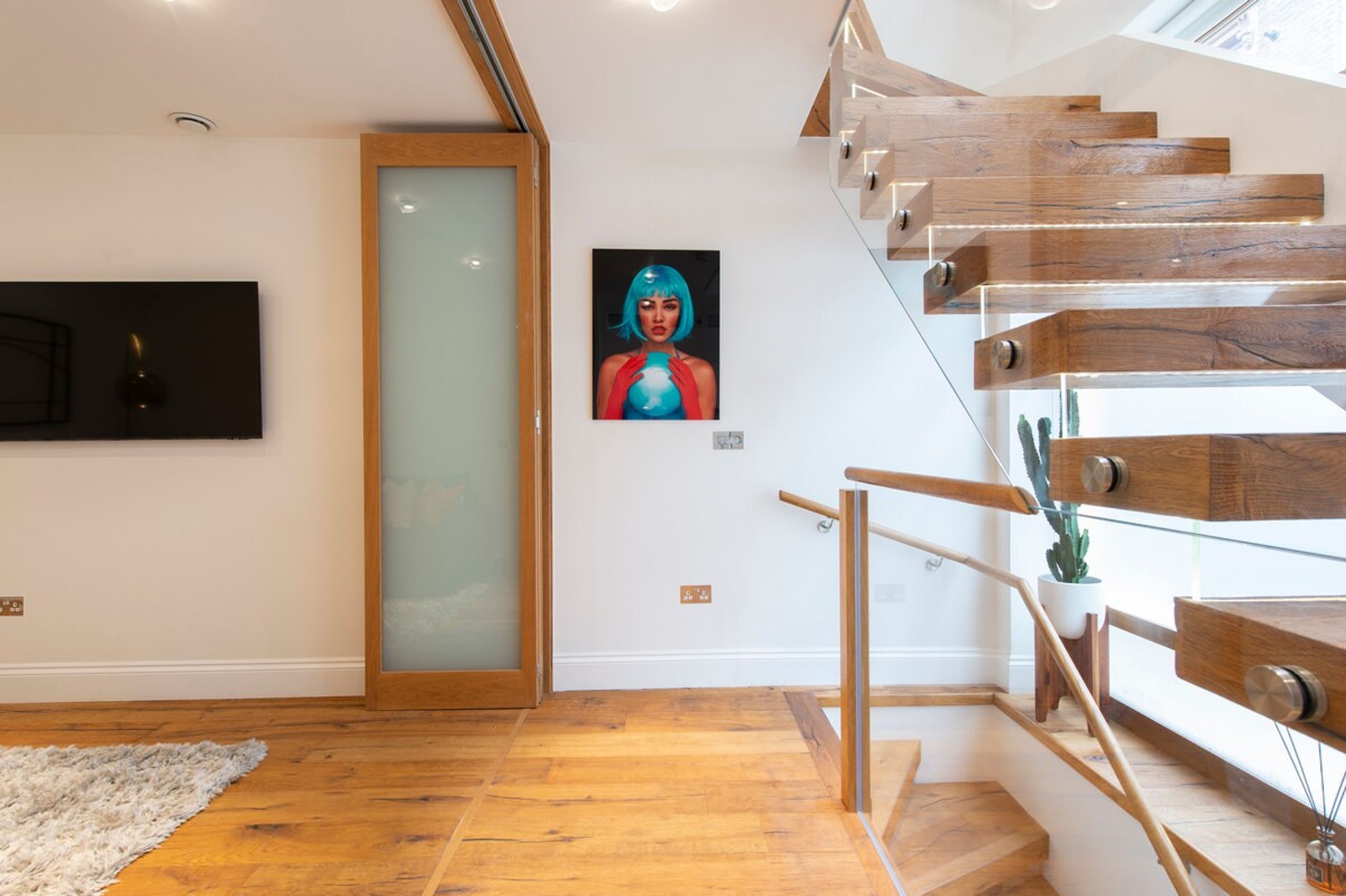 A modern staircase with wooden steps and glass railings, a painting on the wall, and a potted plant.