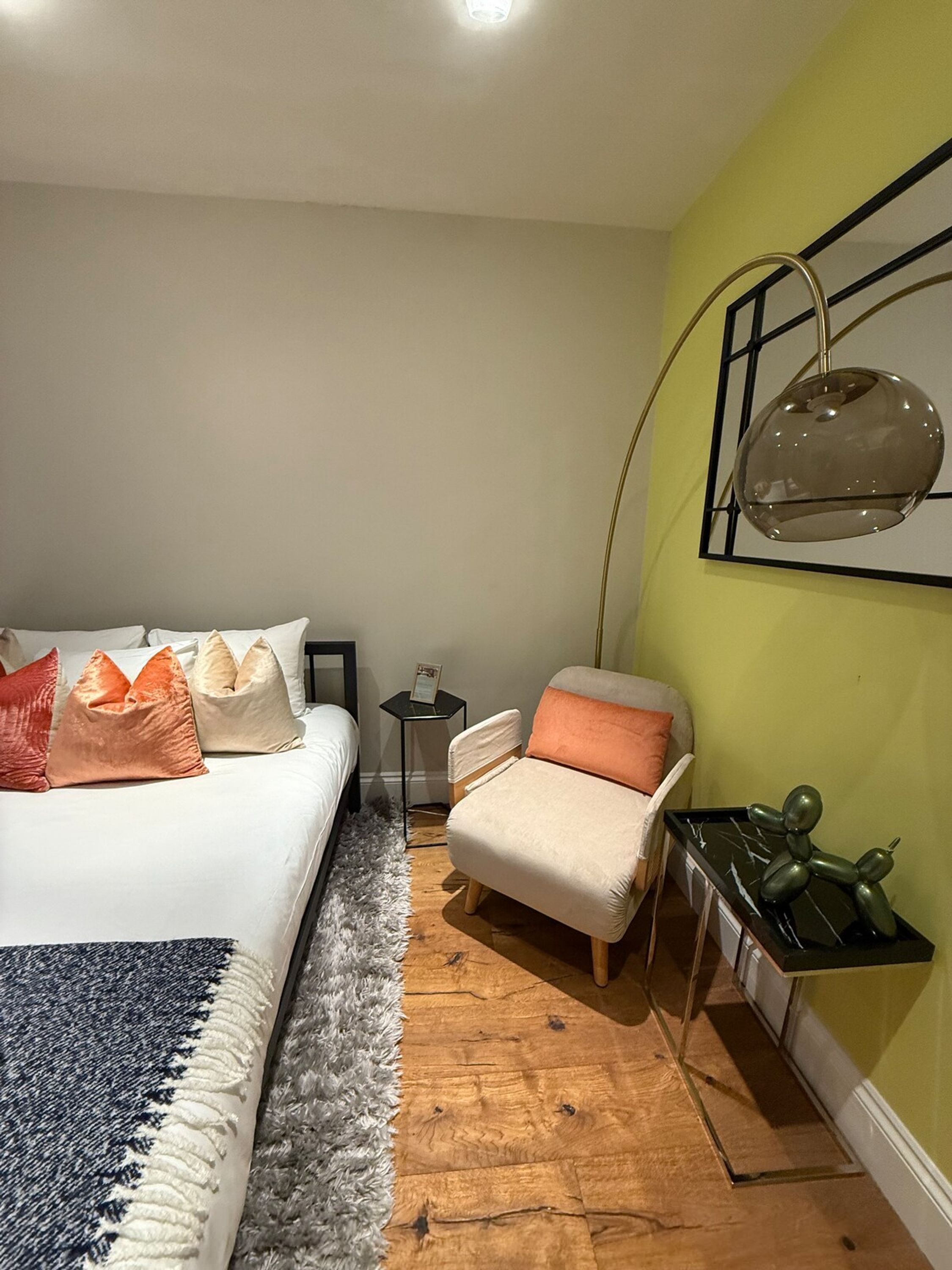 A bedroom with a bed, a chair, a small table, a wall-mounted lamp, and a decorative sculpture.