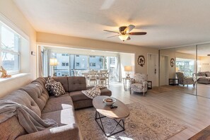 Ein geräumiges Wohnzimmer mit Sofa, Couchtisch und Sesseln, großen Fenstern und einem Deckenventilator.