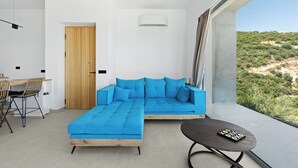 Ein modernes Wohnzimmer mit einer Eckcouch, einem Essbereich mit Stühlen, einem kleinen Tisch und Blick auf eine hügelige Landschaft.