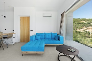 Ein modernes Wohnzimmer mit einer Eckcouch, einem Essbereich mit Stühlen, einem kleinen Tisch und Blick auf eine hügelige Landschaft.