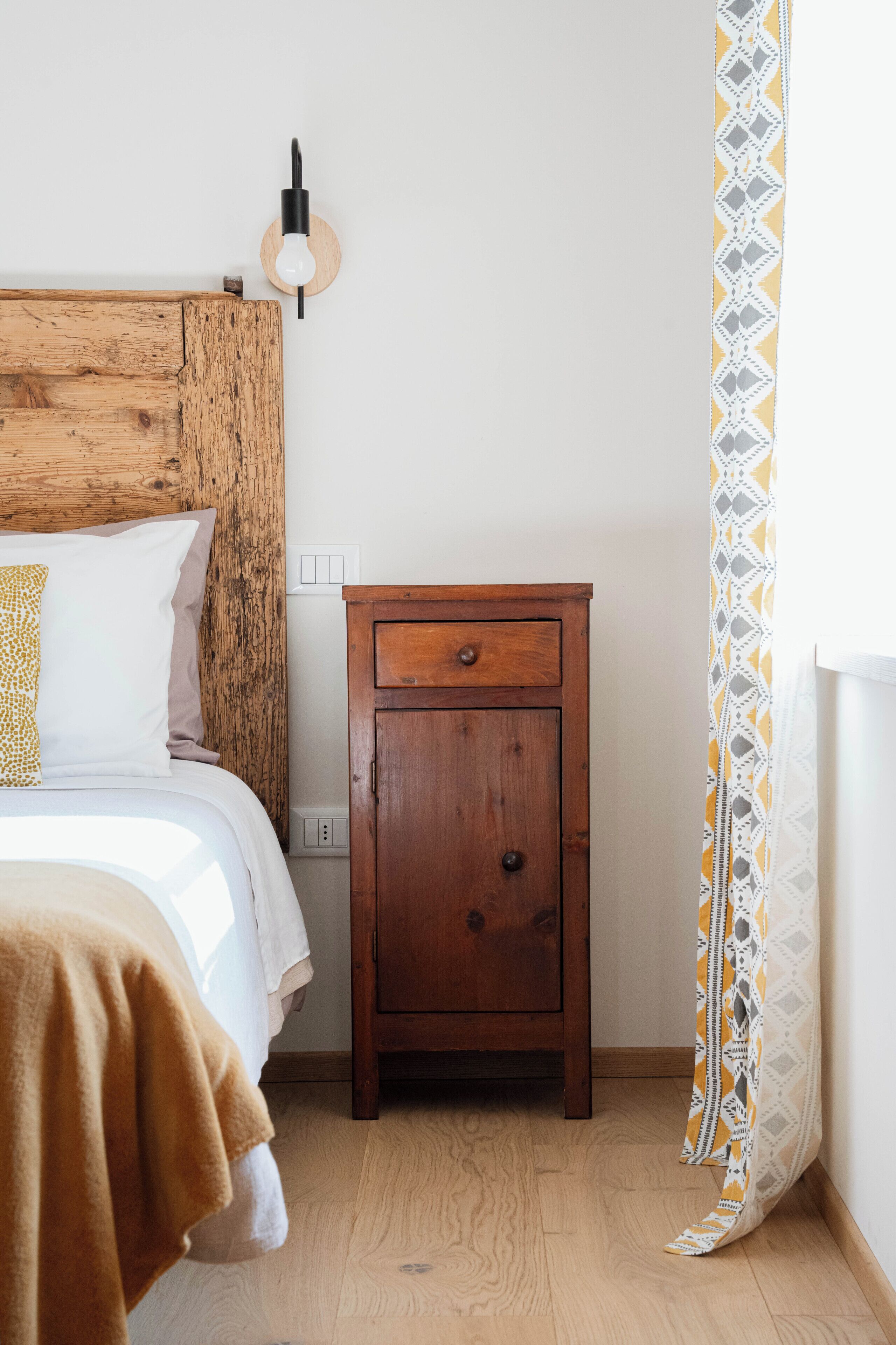 Un dormitorio con cabecera de madera, una mesita de noche de madera con cajón, una cama con almohadas blancas y doradas, y una cortina estampada.