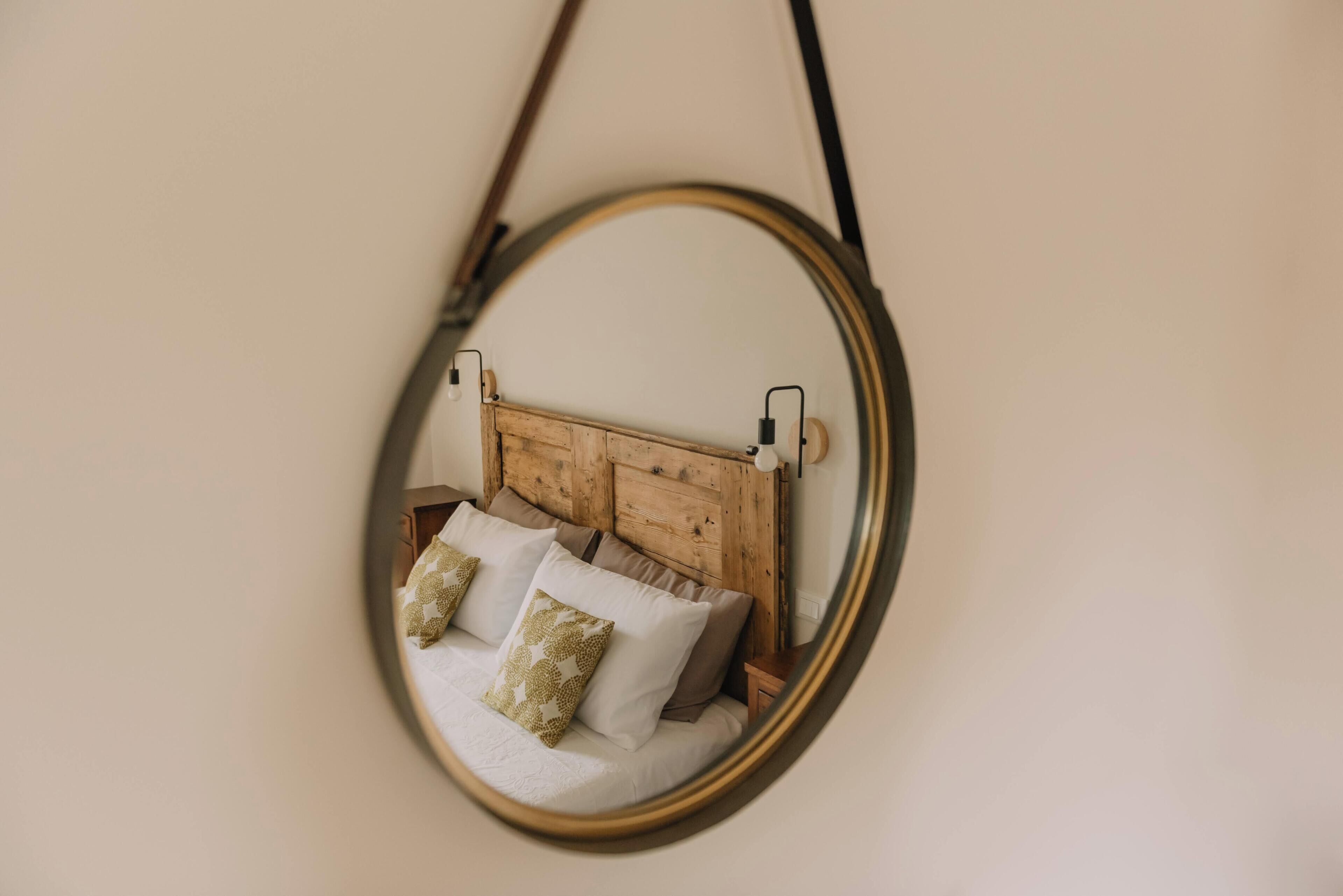 A round mirror with a wooden frame reflecting a bed with pillows.