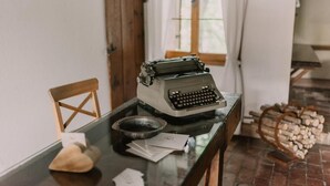 Una máquina de escribir antigua sobre una mesa de vidrio, con papeles, una silla de madera y una ventana con cortinas.