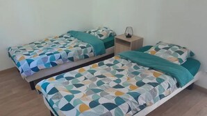 A room with two single beds, each with a patterned bedspread and matching pillows. There is a bedside table with a lamp and a small shelf.