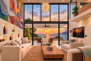 A modern living room with a large window overlooking a sunset, a ceiling fan, and a dining area. - Unique Designer Gem: Private Pool, Elevator, Views (Puerto Vallarta)