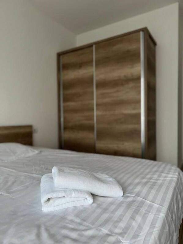 Una cama bien hecha con cubrecama a rayas y toallas blancas dobladas, un armario de madera y una pared lisa.