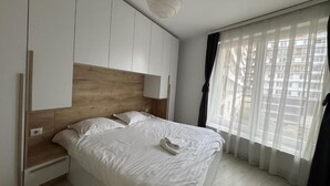 Un dormitorio con una cama, cabecera de madera, mesita de noche, lámpara y ventana con cortinas traslúcidas.