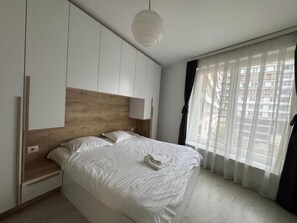Un dormitorio con una cama, cabecera de madera, mesita de noche, lámpara y ventana con cortinas traslúcidas.