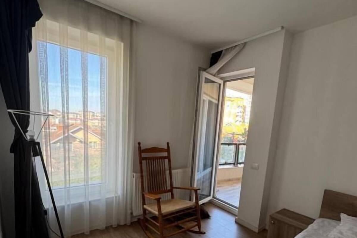A hotel room with a bed, a chair, a lamp, a window with curtains, and a view of buildings outside.
