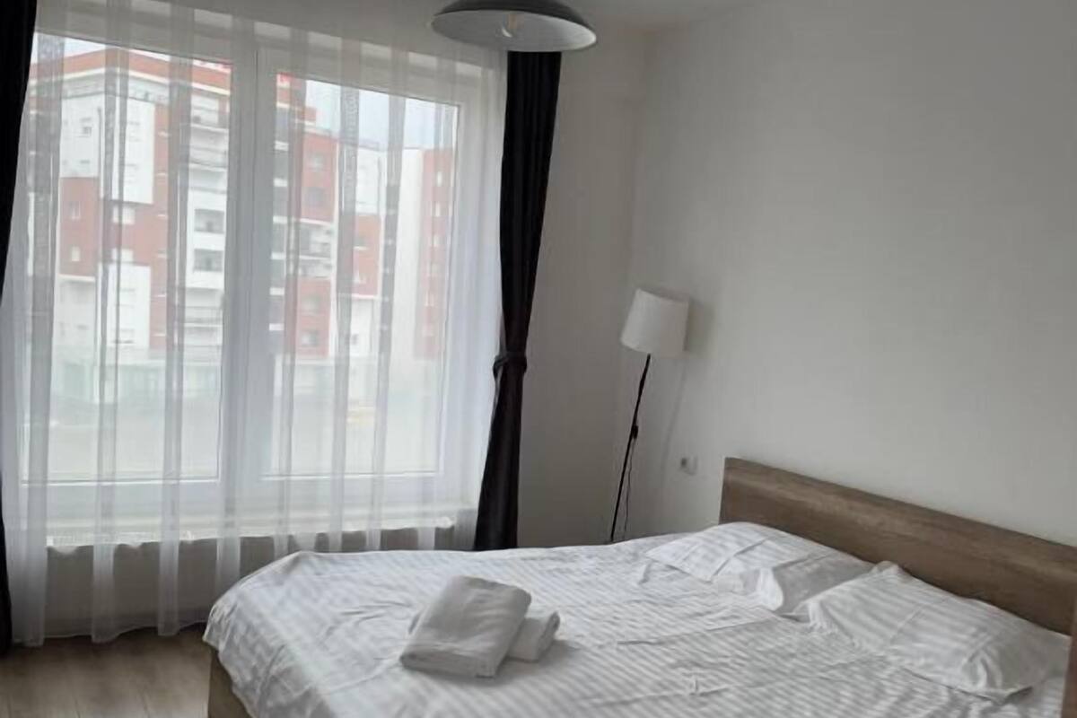 A hotel room with a large bed, a bedside lamp, and a view of buildings through sheer curtains.
