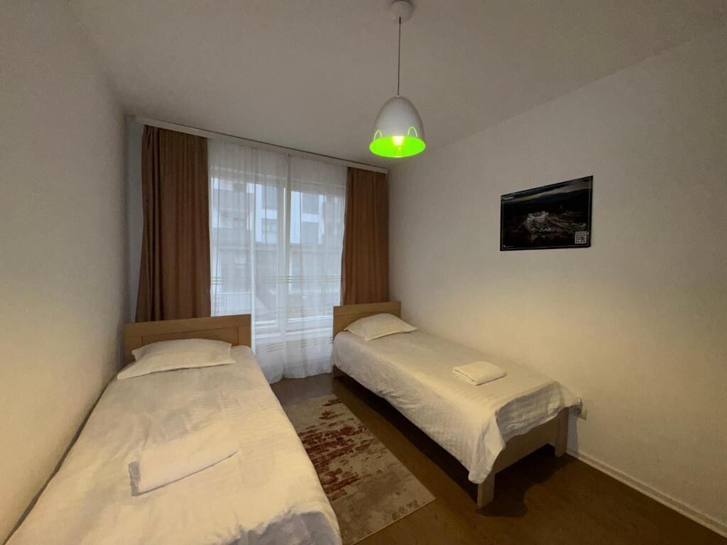A room with two single beds, a window with curtains, a hanging light fixture, and a framed picture on the wall.